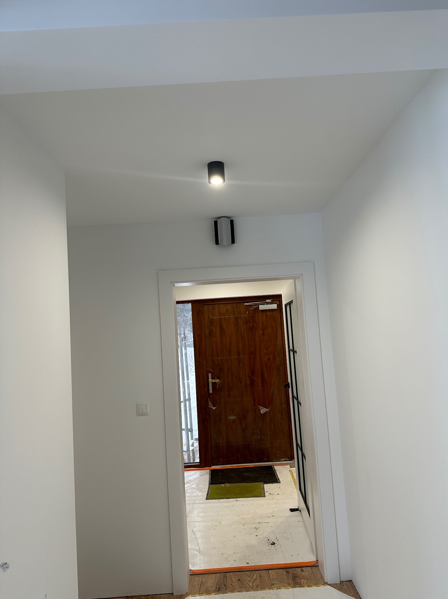 Biały korytarz po remoncie z drewnianymi drzwiami wejściowymi i nowoczesnym oświetleniem sufitowym. Widoczne zabezpieczenia podłogi.