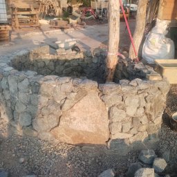 Bartosz Zawada - Kamienna studnia w ogrodzie, zbudowana z nieregularnych bloków skalnych, otoczona żwirem, z widocznymi narzędziami i sprzętem budowlanym w tle.