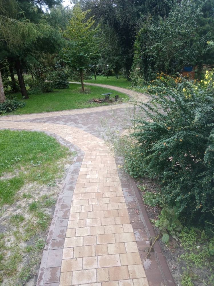 Kaskadowa ścieżka z jasnej kostki brukowej, obrzeża z ciemniejszej kostki. Ścieżka rozwidla się w oddali, otoczona zielenią drzew i krzewów.