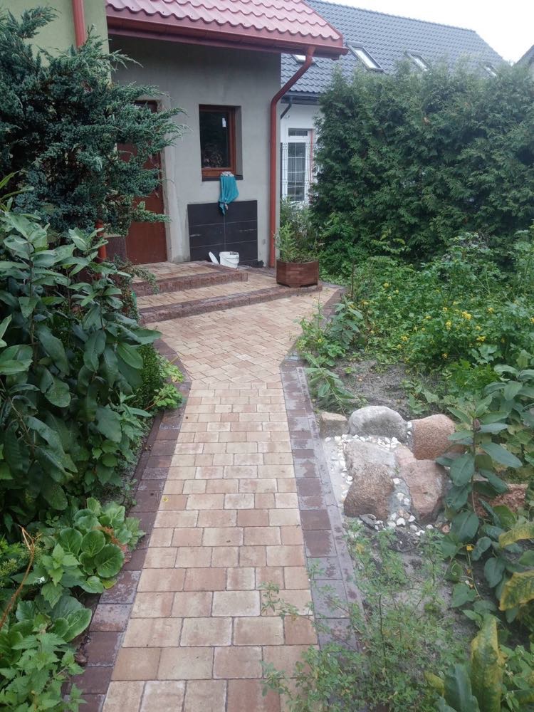 Ułożona kostka brukowa prowadzi do domu z zielonymi nasadzeniami po bokach. Brązowa kostka z ciemniejszym obrzeżem. Widok na wejście i fragment ogrodu.