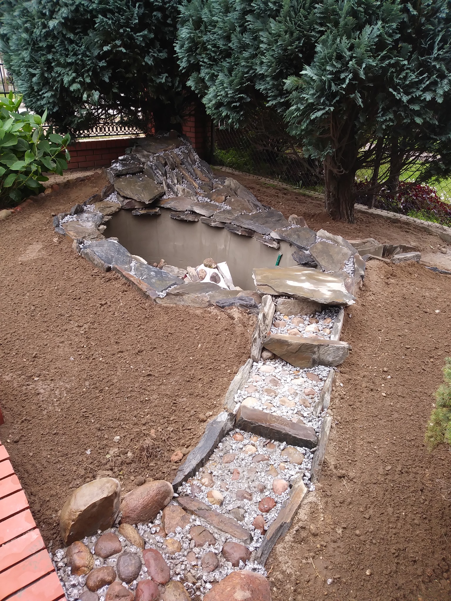 Oczko wodne z kaskadą z kamienia łupanego, otoczone świeżo wyrównaną ziemią. Kamienna ścieżka z drobnymi kamykami prowadzi w dół zbocza, wśród zieleni drzew.