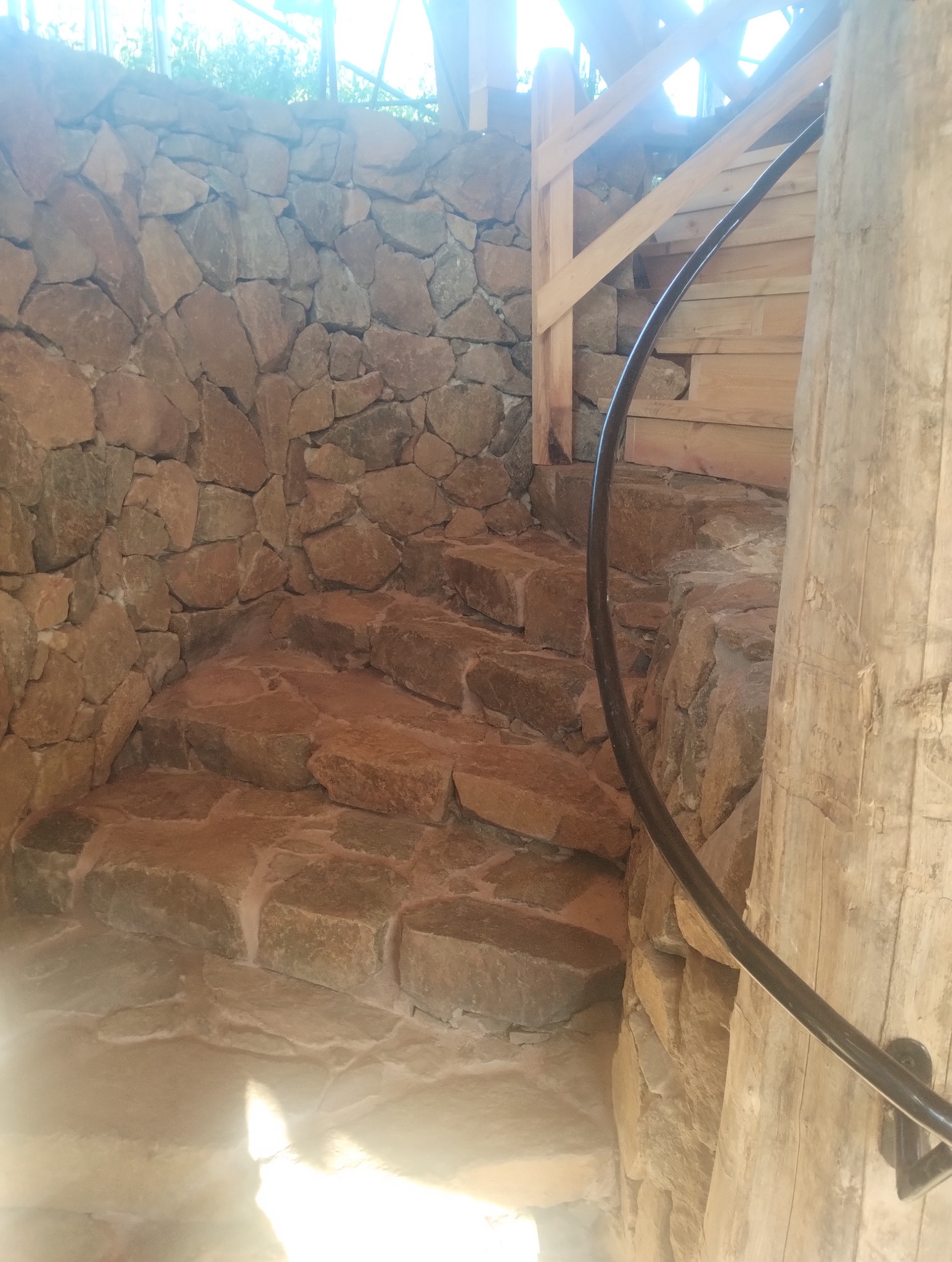 Kamienne schody z drewnianą balustradą i metalową poręczą, prowadzące w górę. Widoczna kamienna ściana i drewniany słup. Oświetlenie słoneczne z góry.