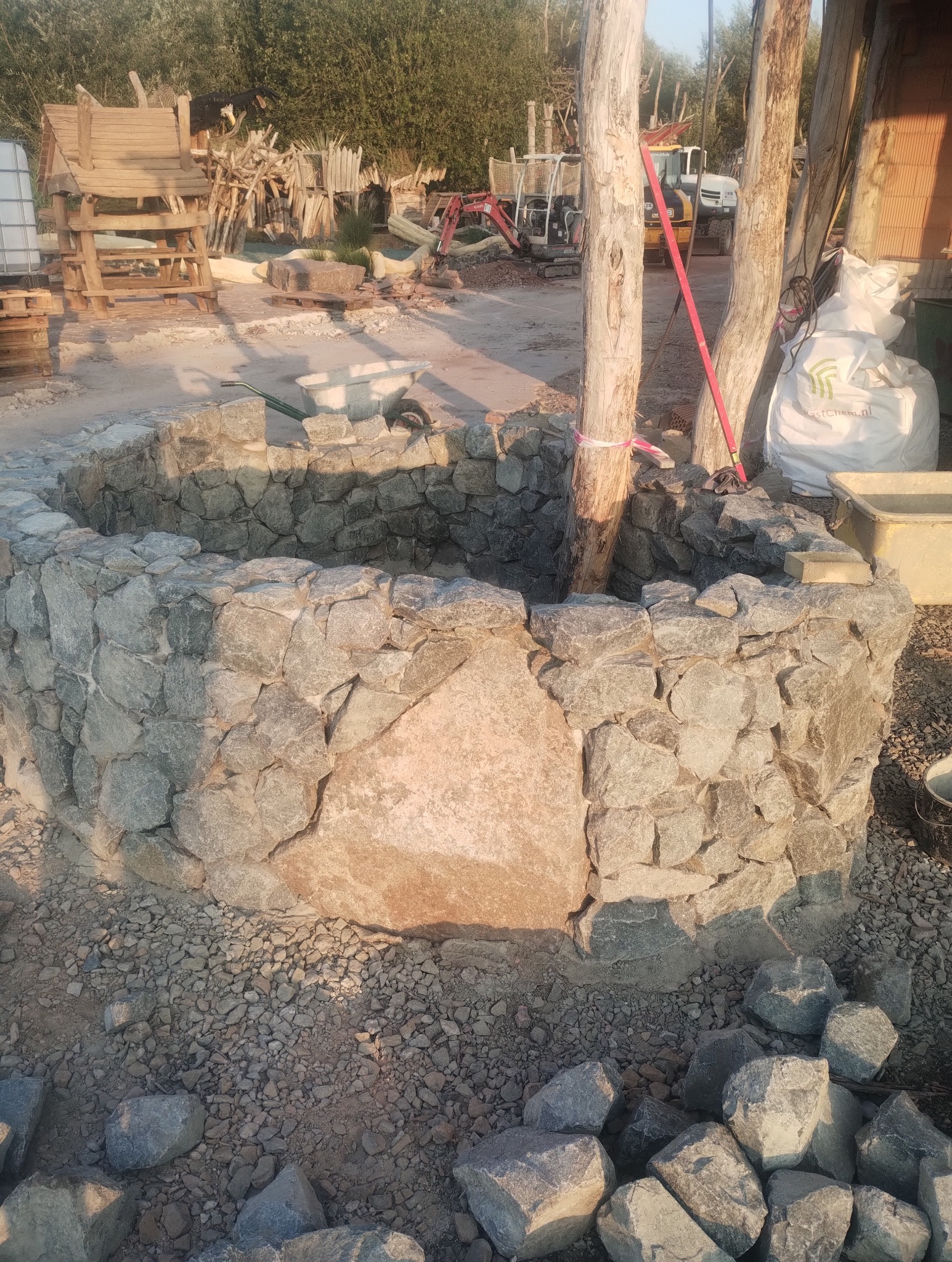 Kamienna studnia w ogrodzie, zbudowana z nieregularnych bloków skalnych, otoczona żwirem, z widocznymi narzędziami i sprzętem budowlanym w tle.