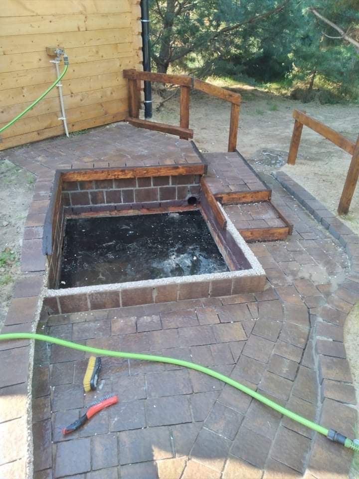 Wykończona strefa relaksu z jacuzzi obudowanym kostką brukową i drewnianymi schodami. Widoczne szczotki i wąż ogrodowy. Całość w rustykalnym stylu.