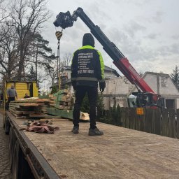 PROFESJONALNE DEKARSTWO - Dekarz w odblaskowej kurtce nadzoruje załadunek drewna dźwigiem Manitou na tle budowy domu. Widoczny numer telefonu firmy dekarskiej na kurtce.