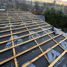 PROFESJONALNE DEKARSTWO - Wysoki kąt na konstrukcję dachu z łatami i membraną w tle krajobrazu. Widoczny fragment buta na dachu. Elementy drewniane w ciepłym odcieniu, membrana czarna z szarym nadrukiem.