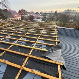 PROFESJONALNE DEKARSTWO - Konstrukcja dachu z drewnianą więźbą i czarną membraną w trakcie montażu. W tle zabudowa jednorodzinna i pochmurne niebo. Widoczny fragment blachy trapezowej.
