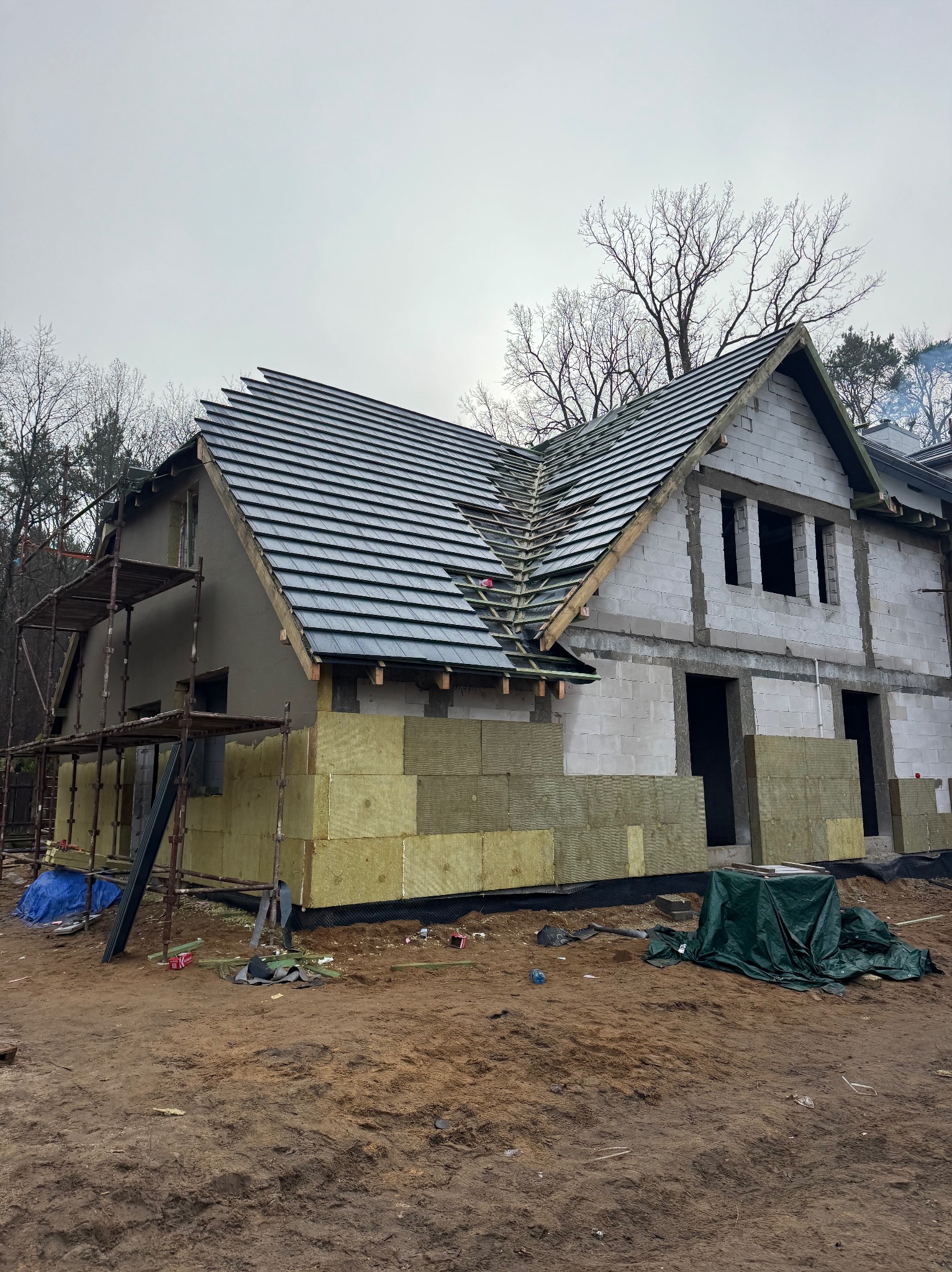Budowa domu jednorodzinnego w trakcie: częściowo położony dach z ciemnej dachówki, ocieplone ściany, widoczne rusztowania i materiały budowlane. Pochmurne niebo w tle.