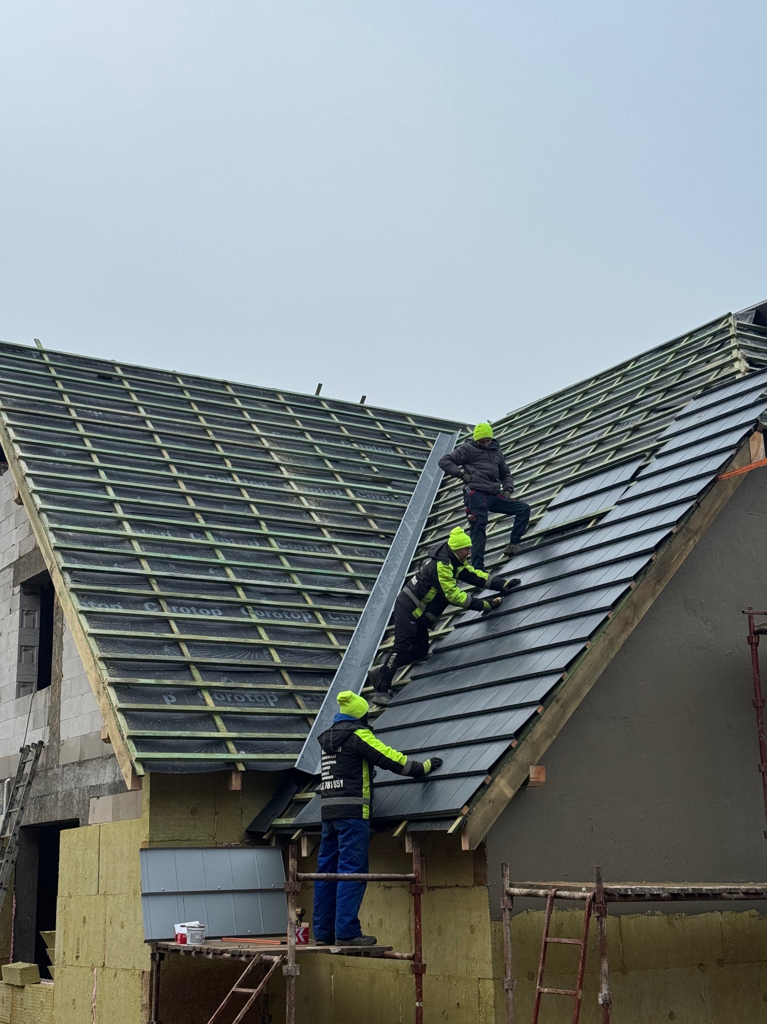 Montaż czarnej dachówki na skośnym dachu domu jednorodzinnego przez trzech dekarzy w odblaskowych ubraniach i zielonych czapkach. Widok z dołu na budowę.