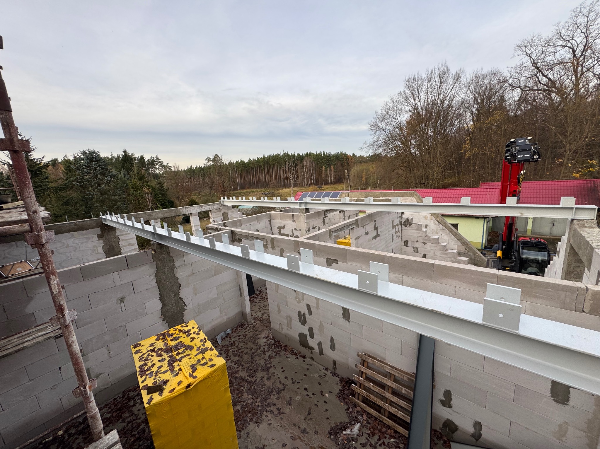 Wznoszenie ścian z bloczków i montaż stalowych belek stropowych na budowie domu jednorodzinnego. Widoczny dźwig i materiały budowlane.