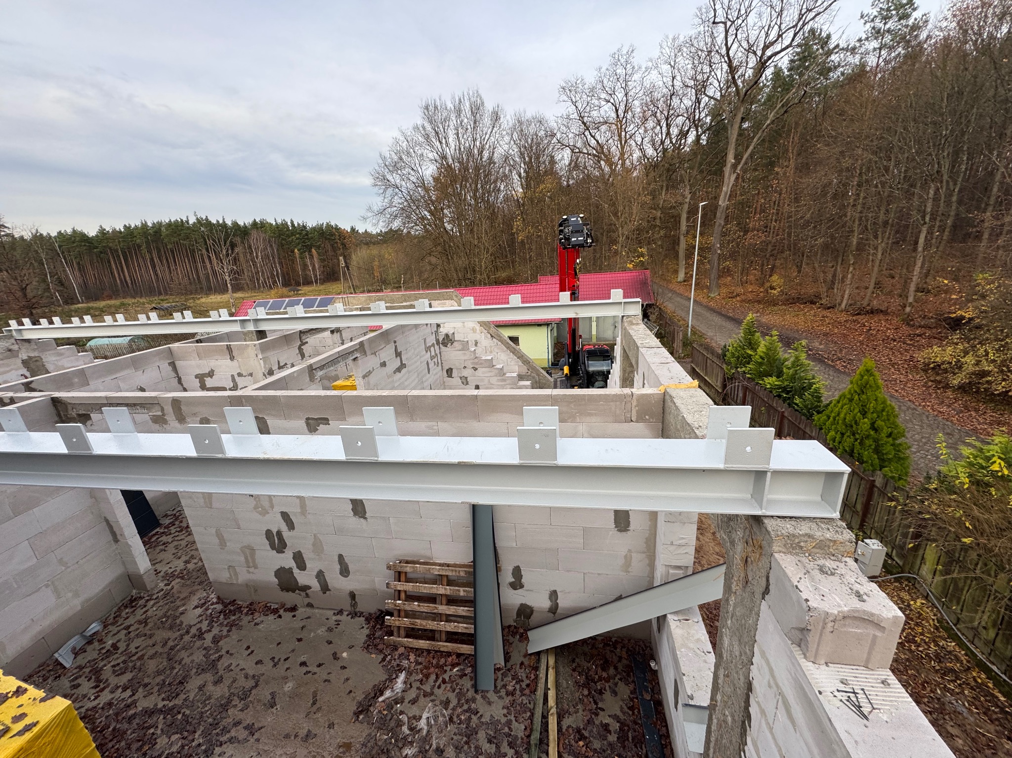 Budowa domu z betonu komórkowego: widok z góry na fundamenty i stalowe belki stropowe, w tle las i czerwony dach sąsiedniego budynku. Etap wznoszenia ścian.