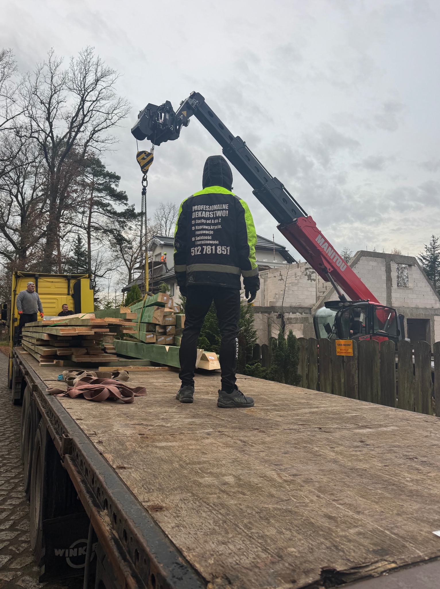 Dekarz w odblaskowej kurtce nadzoruje załadunek drewna dźwigiem Manitou na tle budowy domu. Widoczny numer telefonu firmy dekarskiej na kurtce.
