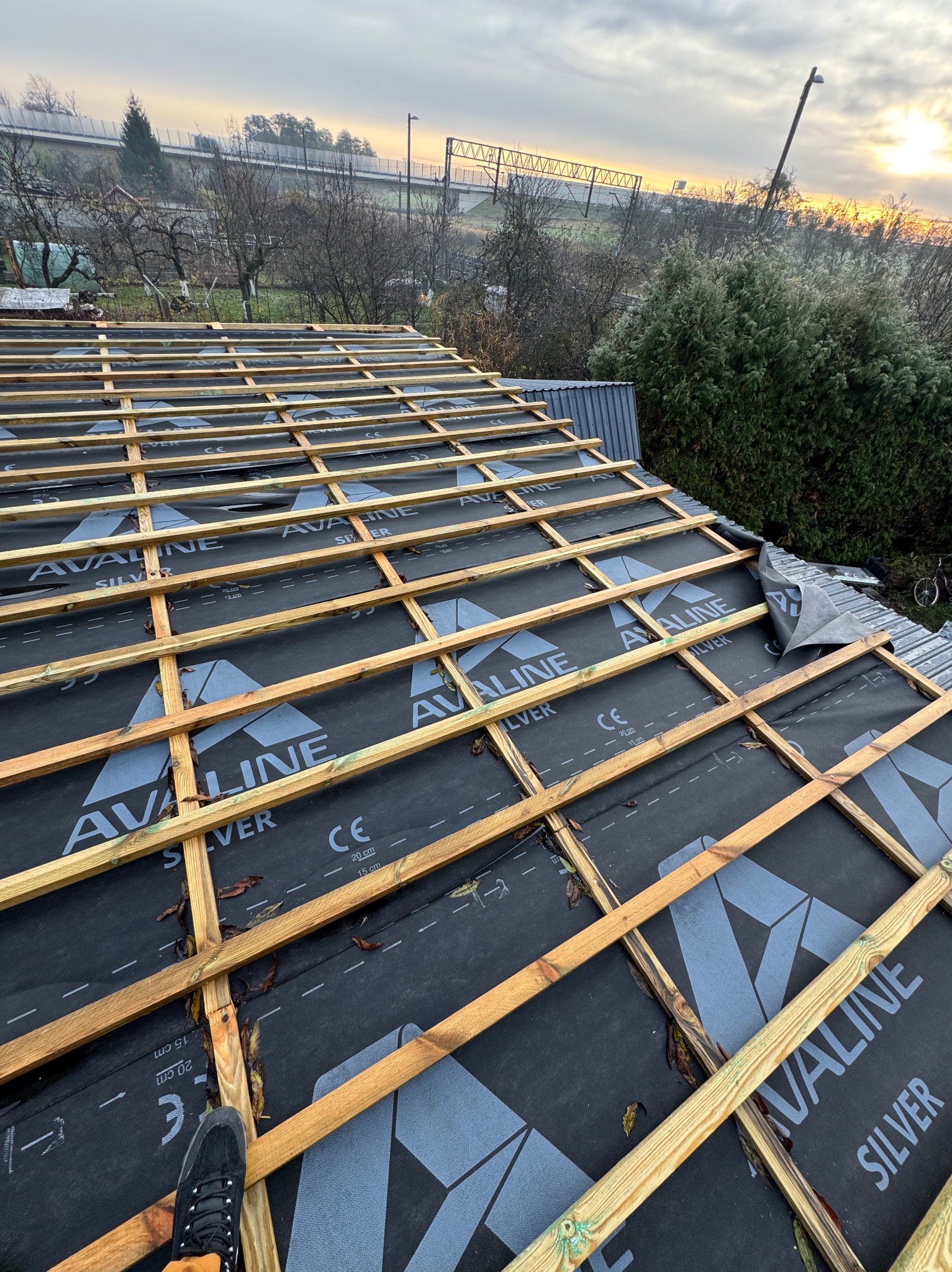 Wysoki kąt na konstrukcję dachu z łatami i membraną w tle krajobrazu. Widoczny fragment buta na dachu. Elementy drewniane w ciepłym odcieniu, membrana czarna z szarym nadrukiem.