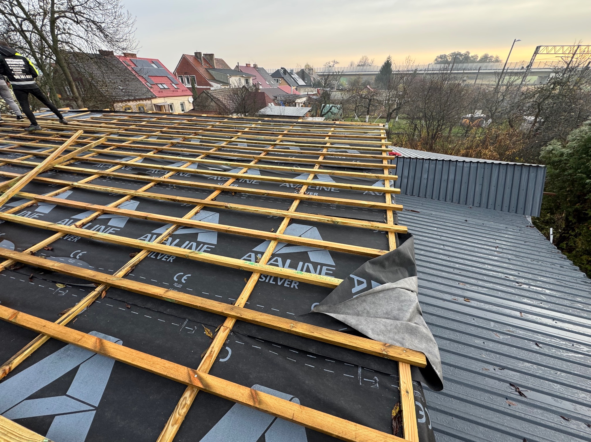 Konstrukcja dachu z drewnianą więźbą i czarną membraną w trakcie montażu. W tle zabudowa jednorodzinna i pochmurne niebo. Widoczny fragment blachy trapezowej.