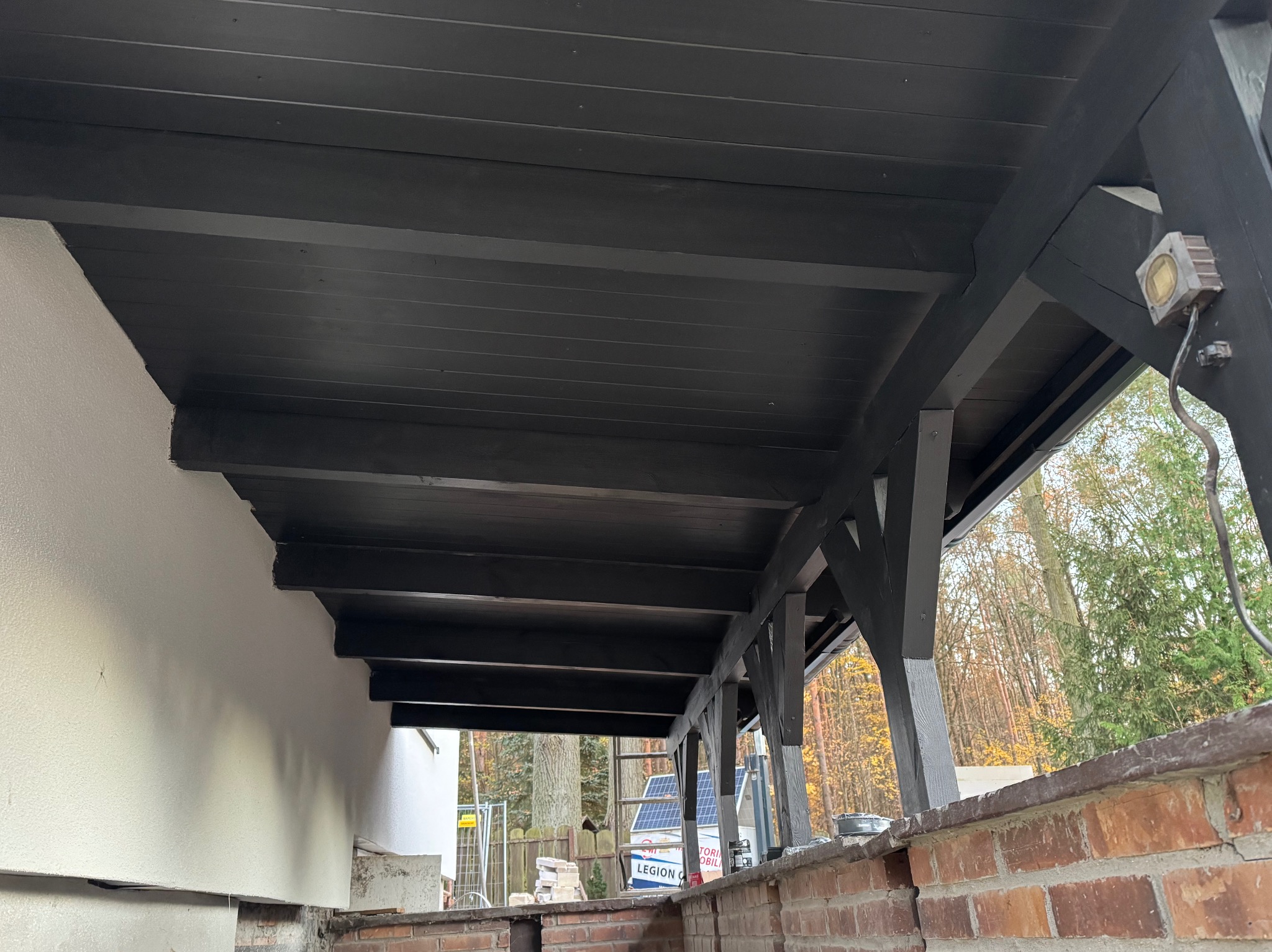 Ciemna, drewniana konstrukcja z oświetleniem zewnętrznym, widok z dołu. Elementy konstrukcyjne pomalowane na czarno, w tle drzewa i fragment budynku.