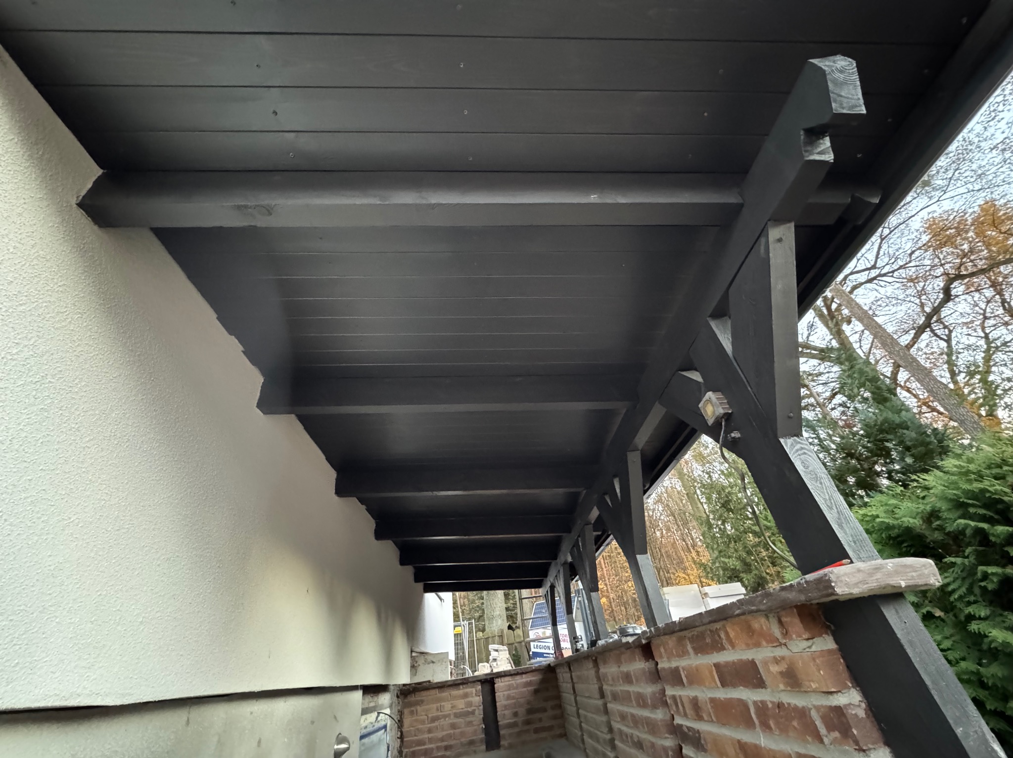 Ciemna, drewniana konstrukcja z belek, prawdopodobnie zadaszenie tarasu lub werandy, z widoczną ścianą budynku i fragmentem ceglanej podstawy. Widok z dołu.