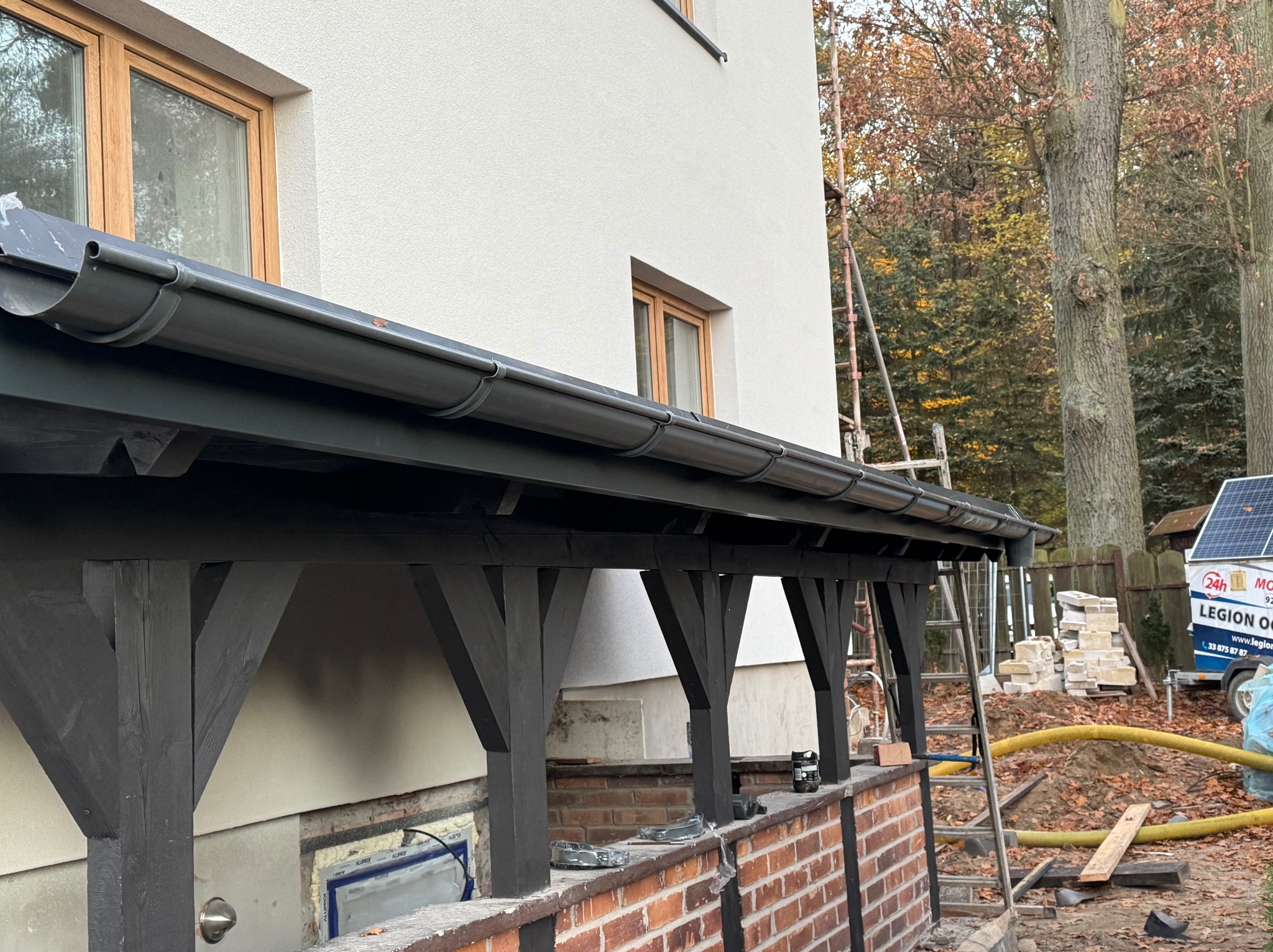 Szare rynny zamontowane na drewnianej konstrukcji przy białym budynku z oknami. W tle drzewa i przyczepka z panelami słonecznymi.