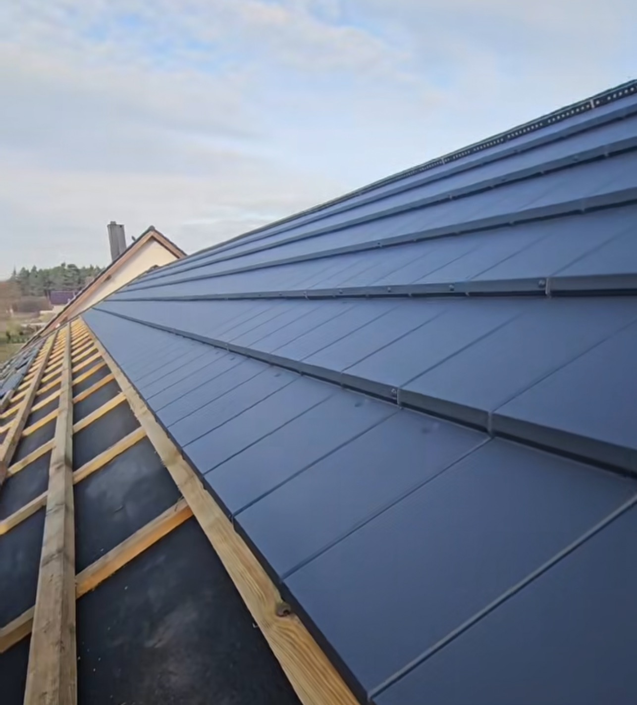 Nowy, grafitowy dach z blachy trapezowej w trakcie montażu, widok z dołu na drewnianą konstrukcję i częściowo pokrytą powierzchnię. Ujęcie pod kątem.