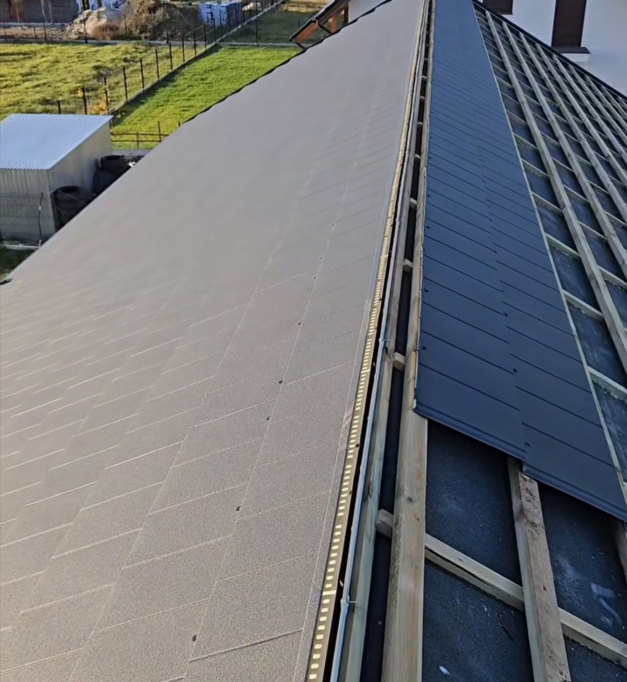 Częściowo pokryty dach z widoczną konstrukcją drewnianą i fragmentem gotowego pokrycia z szarej dachówki bitumicznej oraz czarnej blachy na rąbek stojący.