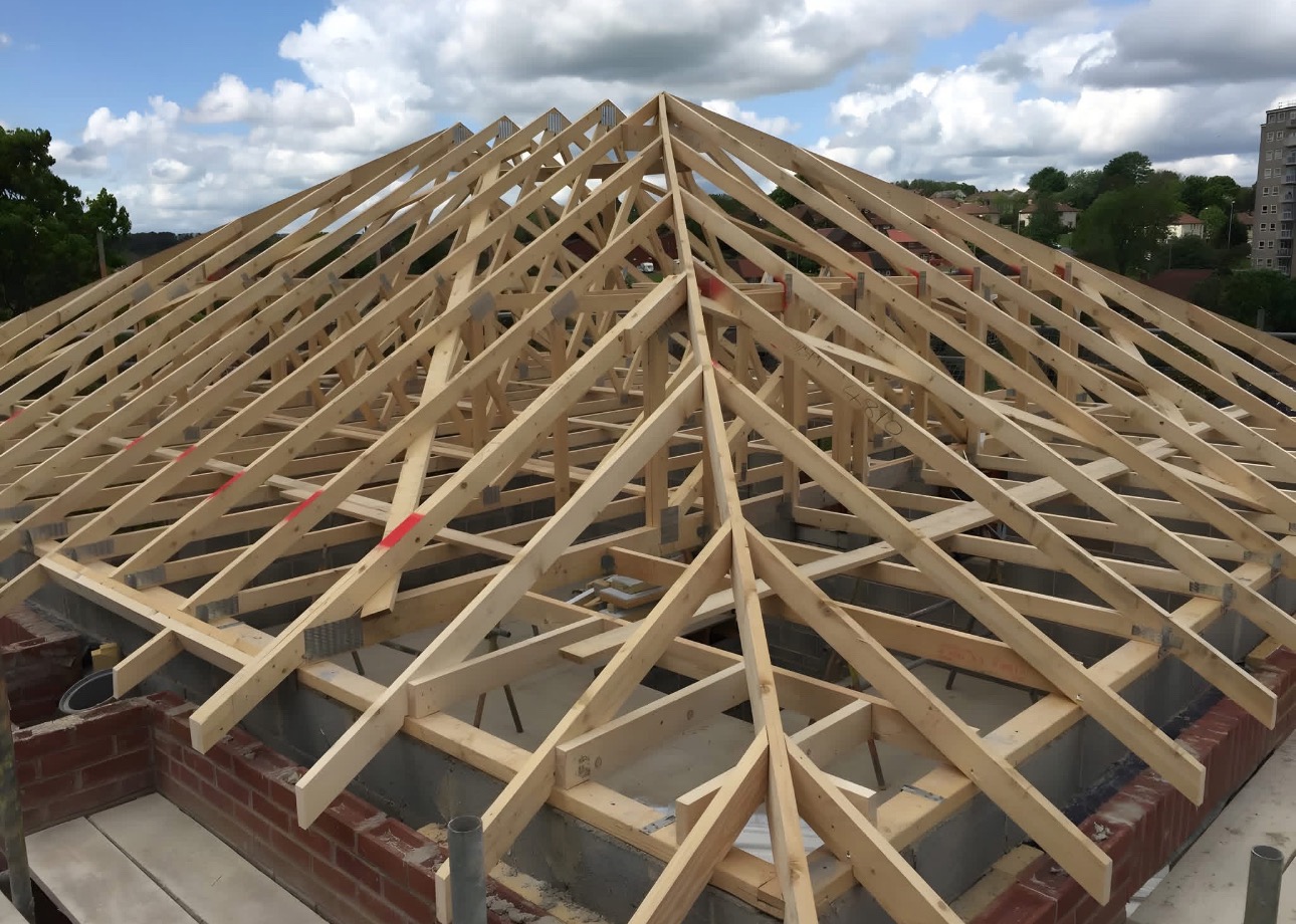 Wznoszenie drewnianej konstrukcji dachu z widocznymi elementami więźby, na tle błękitnego nieba z chmurami i zabudowań w oddali. Widok z góry na surową konstrukcję.