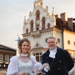 Konrad Barowicz - Para w strojach historycznych na tle ratusza z zegarem. Kobieta w jasnej sukni z koronką, mężczyzna w granatowym fraku i białej koszuli. Sesja plenerowa.