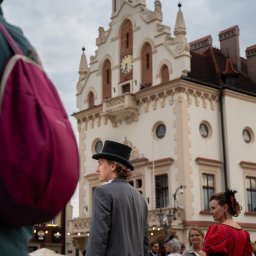 Konrad Barowicz - Sesja plenerowa: mężczyzna w cylindrze i kobieta w czerwonej sukni epokowej na tle zabytkowego ratusza z zegarem. Ujęcie z tłumem w tle.