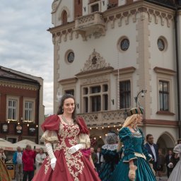 Konrad Barowicz - Uczestniczki sesji w barwnych, historycznych sukniach na tle budynku z zegarem. Detal: bogate zdobienia sukni, białe rękawiczki, elegancka biżuteria. Sesja plenerowa.