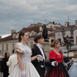 Konrad Barowicz - Grupa osób w stylizowanych, historycznych strojach spaceruje po placu miejskim. Kobiety w sukniach balowych, mężczyzna w smokingu. Tło stanowi architektura miasta.