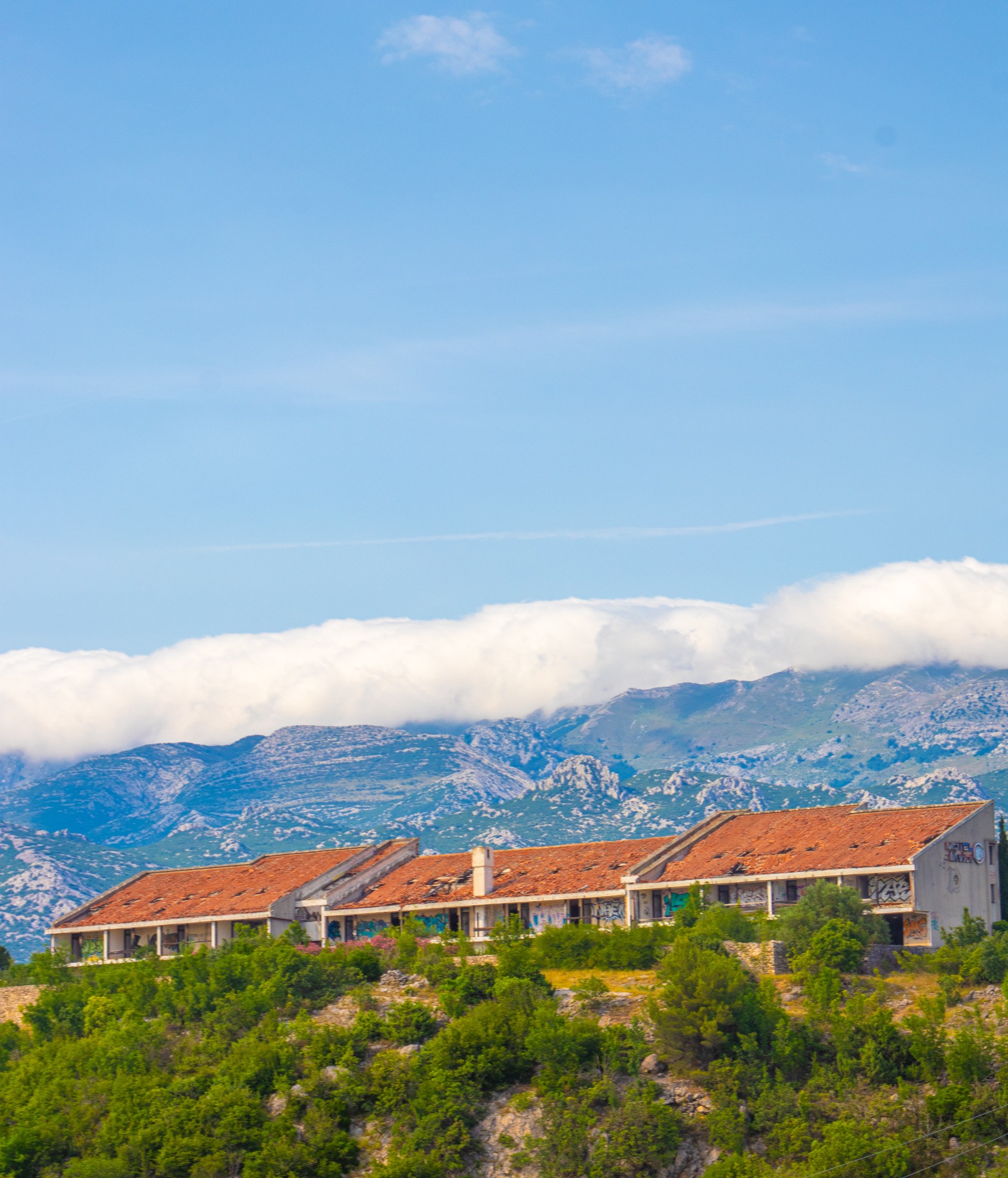 Opuszczony budynek z czerwoną dachówką na wzgórzu, z graffiti na ścianach, na tle gór i błękitnego nieba z chmurami. Krajobraz z zaniedbaną architekturą.