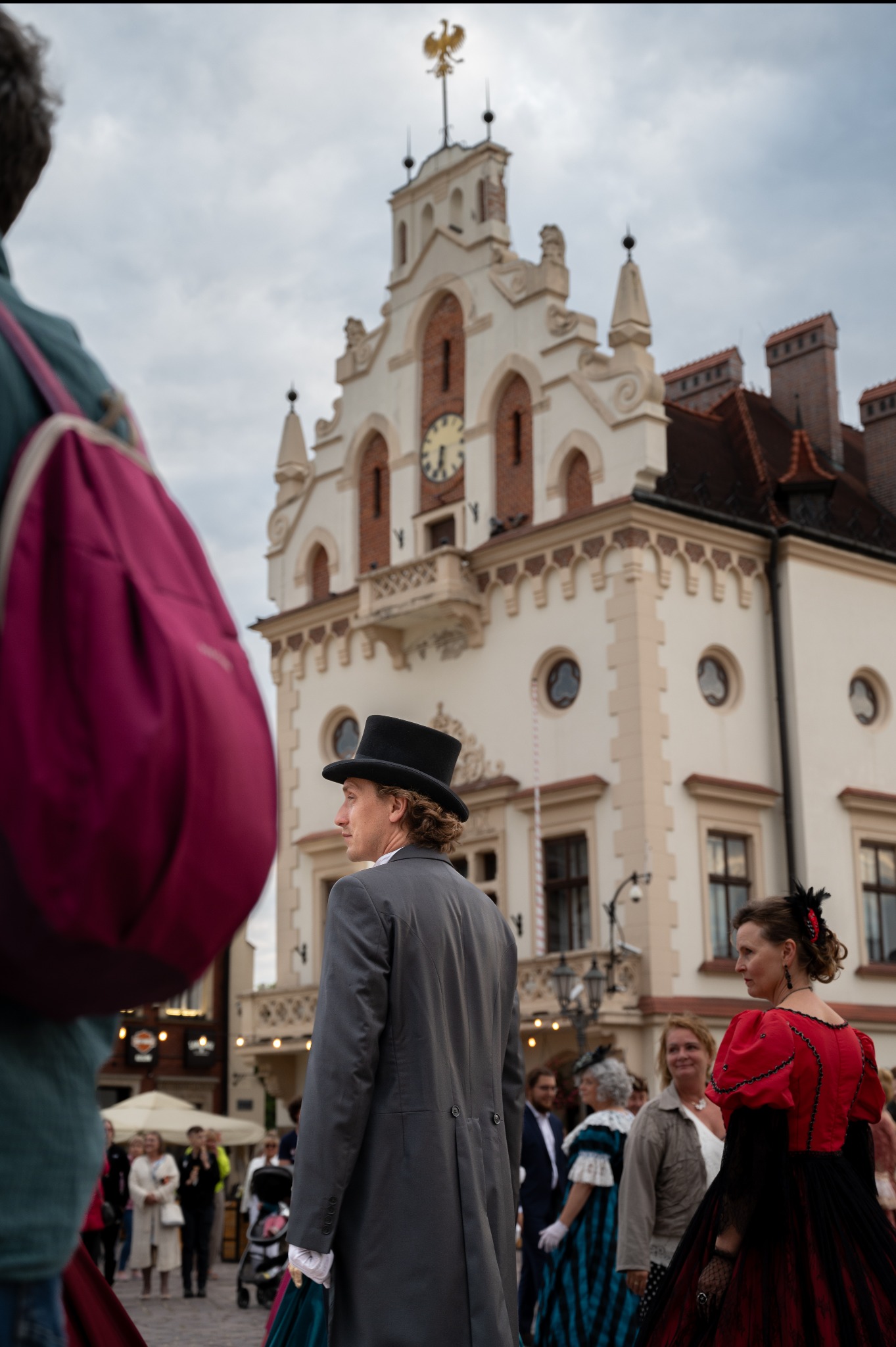 Sesja plenerowa: mężczyzna w cylindrze i kobieta w czerwonej sukni epokowej na tle zabytkowego ratusza z zegarem. Ujęcie z tłumem w tle.