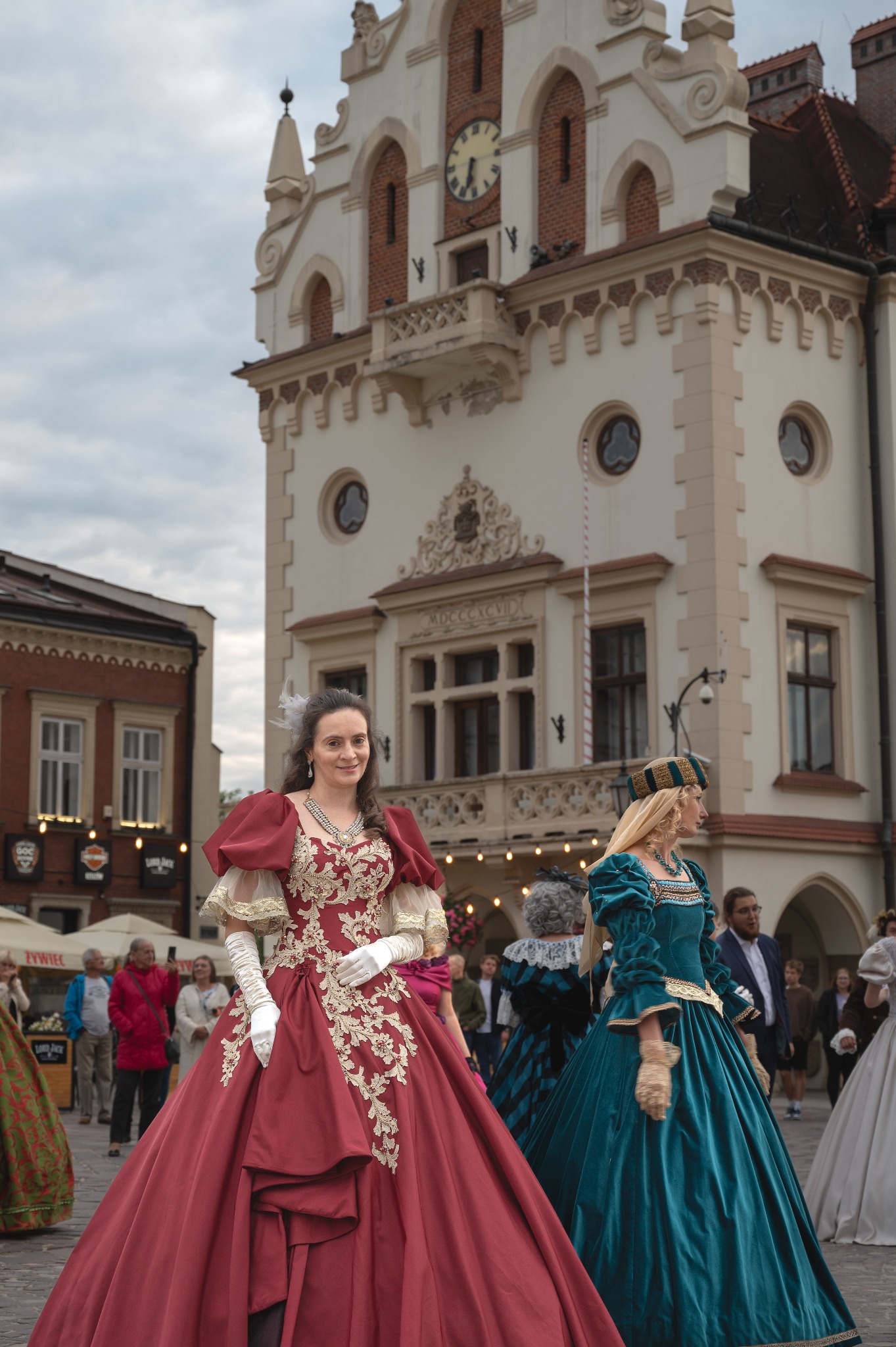 Uczestniczki sesji w barwnych, historycznych sukniach na tle budynku z zegarem. Detal: bogate zdobienia sukni, białe rękawiczki, elegancka biżuteria. Sesja plenerowa.