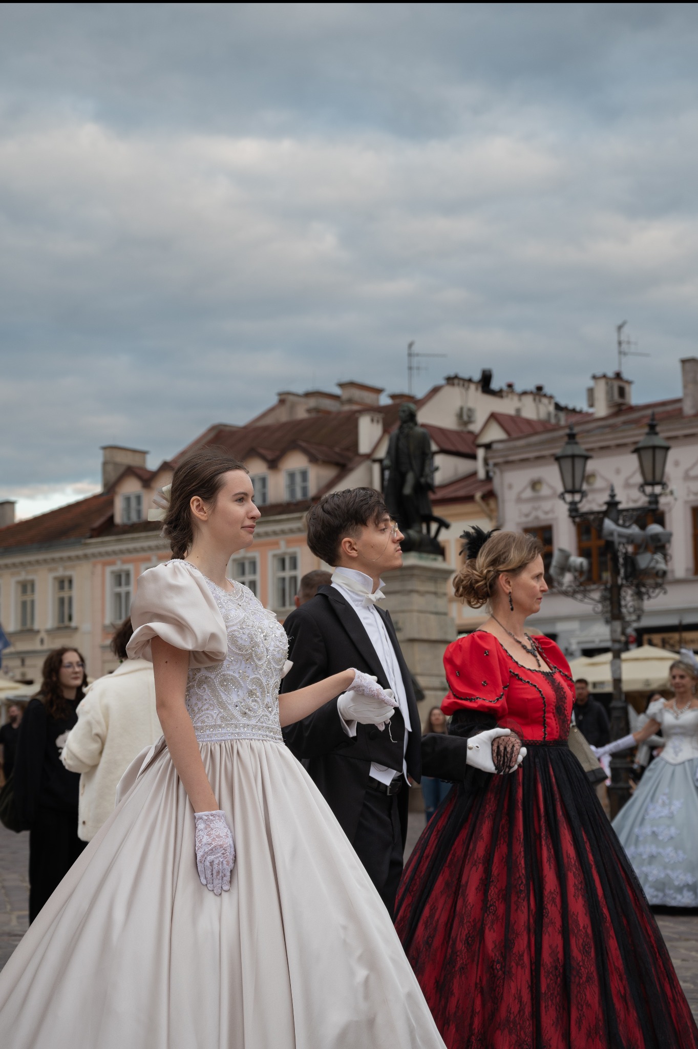 Grupa osób w stylizowanych, historycznych strojach spaceruje po placu miejskim. Kobiety w sukniach balowych, mężczyzna w smokingu. Tło stanowi architektura miasta.