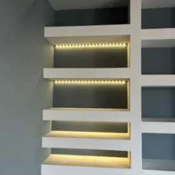 Białe, minimalistyczne półki z oświetleniem LED na szaro-niebieskiej ścianie. Widoczny fragment grzejnika pod dolną półką. Geometryczny układ półek.