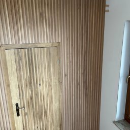 Usługi Ogólnobudowlane Mania Sebastian - Drewniana boazeria lamelowa na ścianie, zintegrowana z drzwiami, tworząca spójny wizualnie panel. Widoczne elementy montażowe u góry ściany.