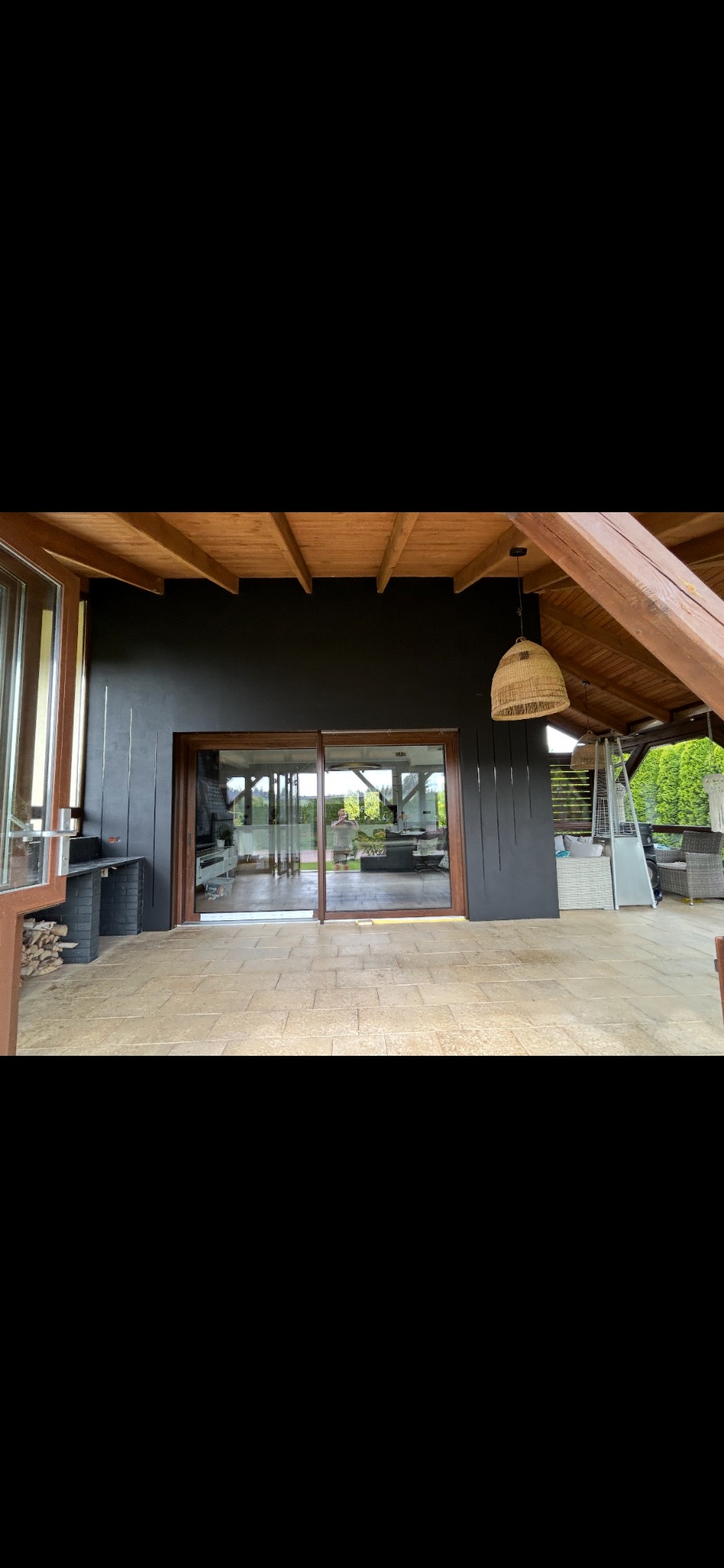 Zadaszony taras z drewnianą konstrukcją i dużymi przesuwnymi oknami. Ściana pomalowana na ciemny kolor, podłoga wyłożona płytkami. Widoczne elementy wyposażenia.