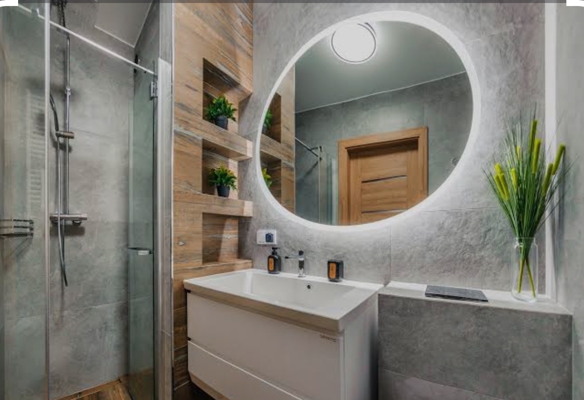 Nowoczesna łazienka z kabiną prysznicową, umywalką z szafką i podświetlanym lustrem owalnym. Na ścianie drewniane półki z roślinami.