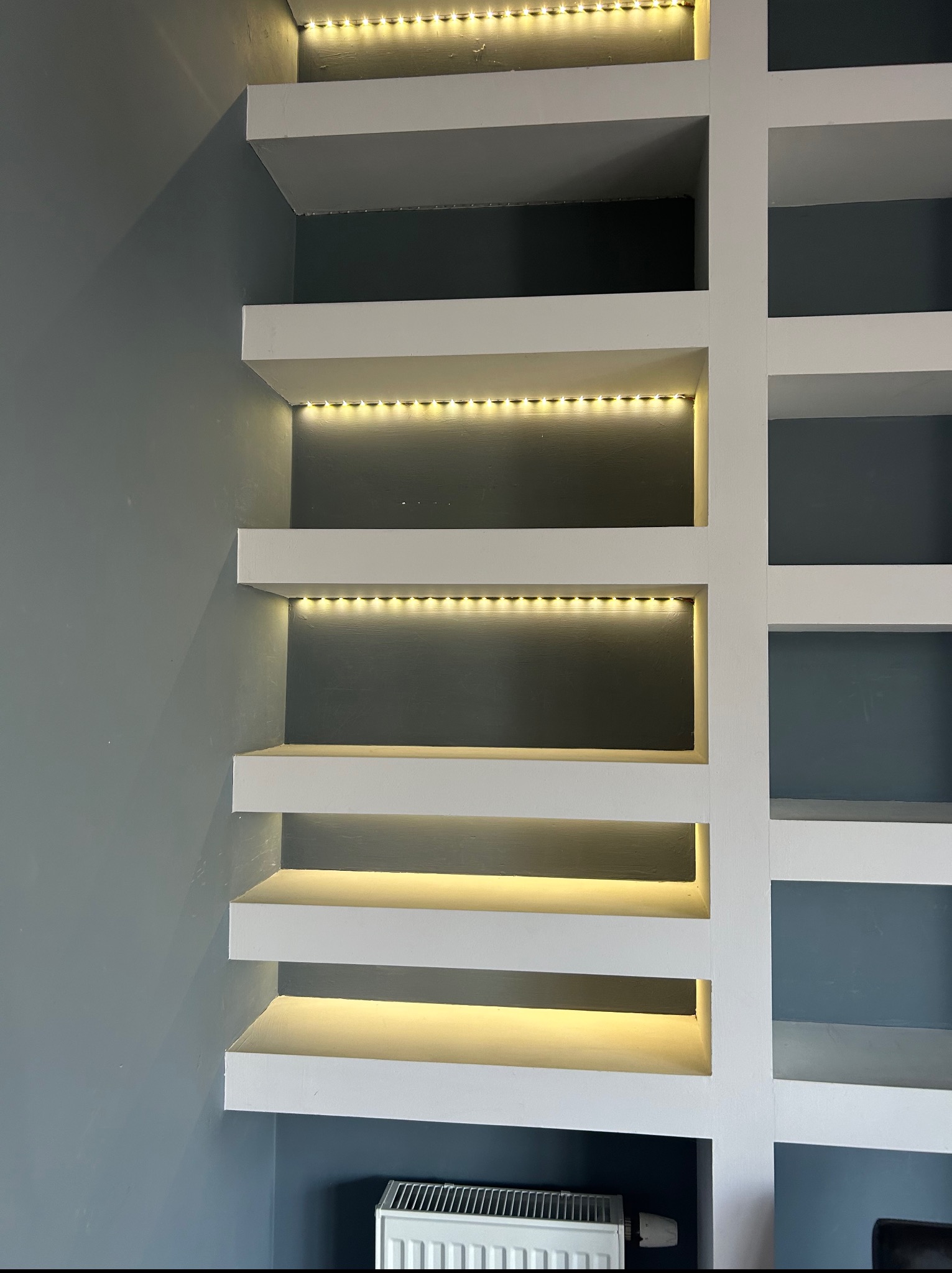 Białe, minimalistyczne półki z oświetleniem LED na szaro-niebieskiej ścianie. Widoczny fragment grzejnika pod dolną półką. Geometryczny układ półek.