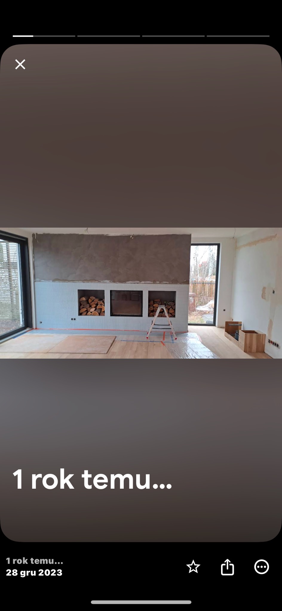 Wnętrze w trakcie prac wykończeniowych: szpachlowana ściana nad kominkiem z wnękami na drewno, podłoga zabezpieczona folią, drabina. Widok na okno i drzewa na zewnątrz.