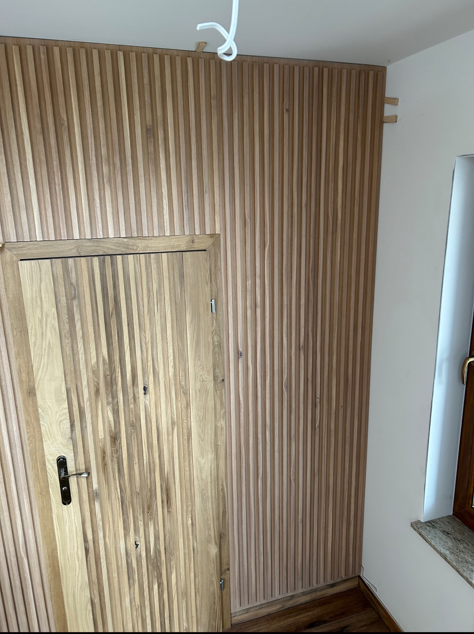 Drewniana boazeria lamelowa na ścianie, zintegrowana z drzwiami, tworząca spójny wizualnie panel. Widoczne elementy montażowe u góry ściany.