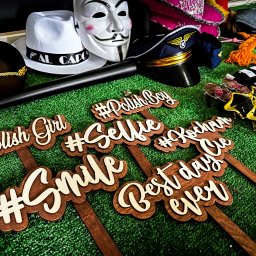 Firma Handlowo-Usługowa Wojciech Peszke - Różnorodne rekwizyty do fotobudki: maska, kapelusze, okulary i drewniane tabliczki z napisami #Selfie, #Smile, Best day ever na zielonym tle.