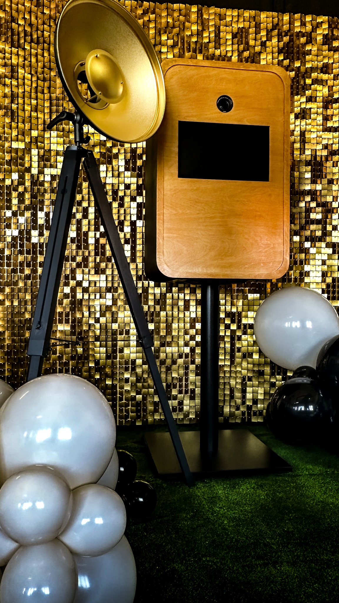 Drewniana fotobudka z lampą na trójnogu, na tle złotej ściany i balonów. Dekoracja eventowa z zieloną trawą jako podłożem.