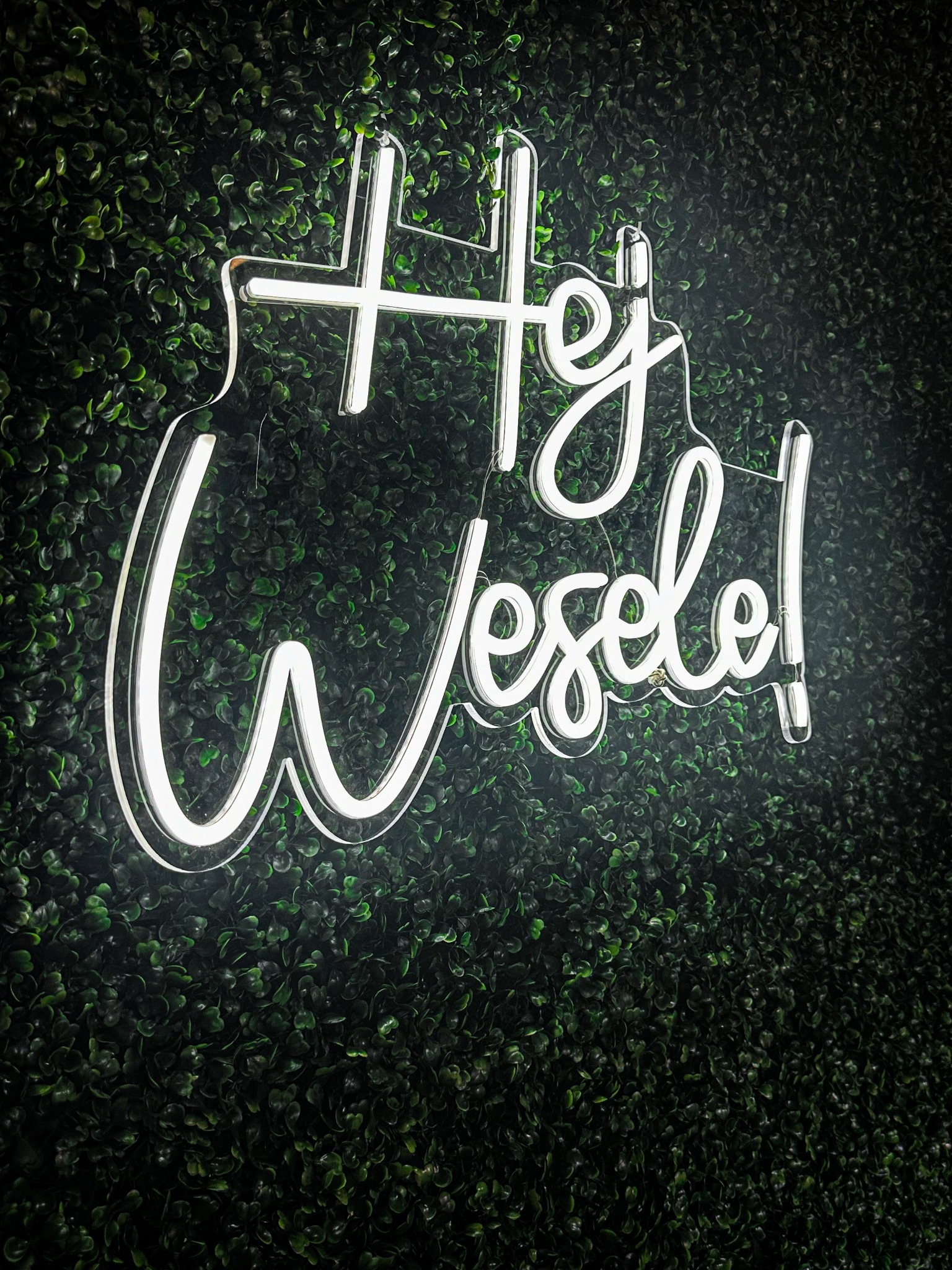 Świecący neon 'Hej Wesele!' na tle ciemnozielonej ściany z roślin. Dekoracja sali weselnej, nowoczesny akcent świetlny. Idealne do fotobudki.