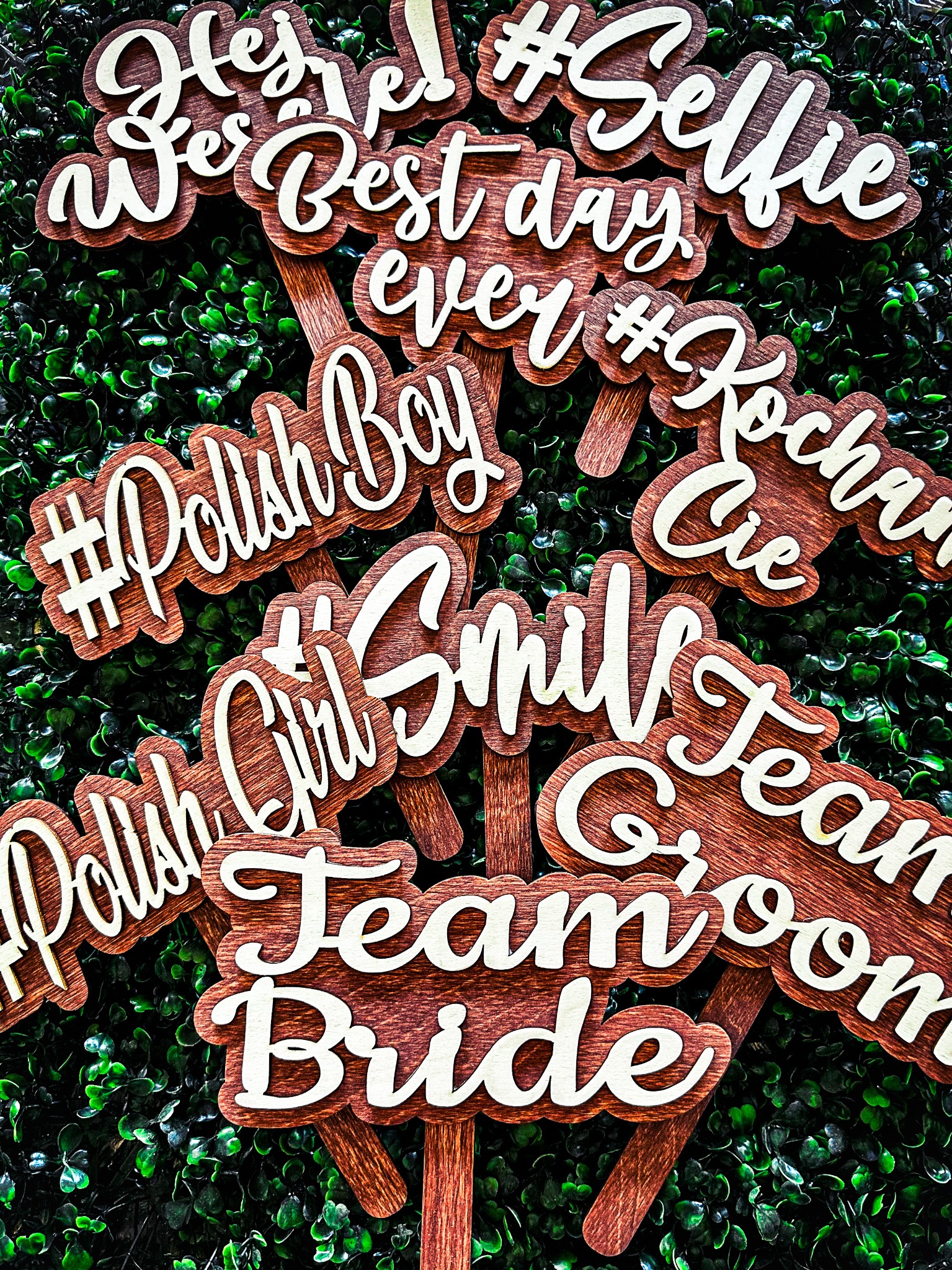 Drewniane rekwizyty do fotobudki z napisami: #Selfie, Best Day Ever, #PolishBoy, Team Bride, na tle zielonej ściany z drobnych liści.