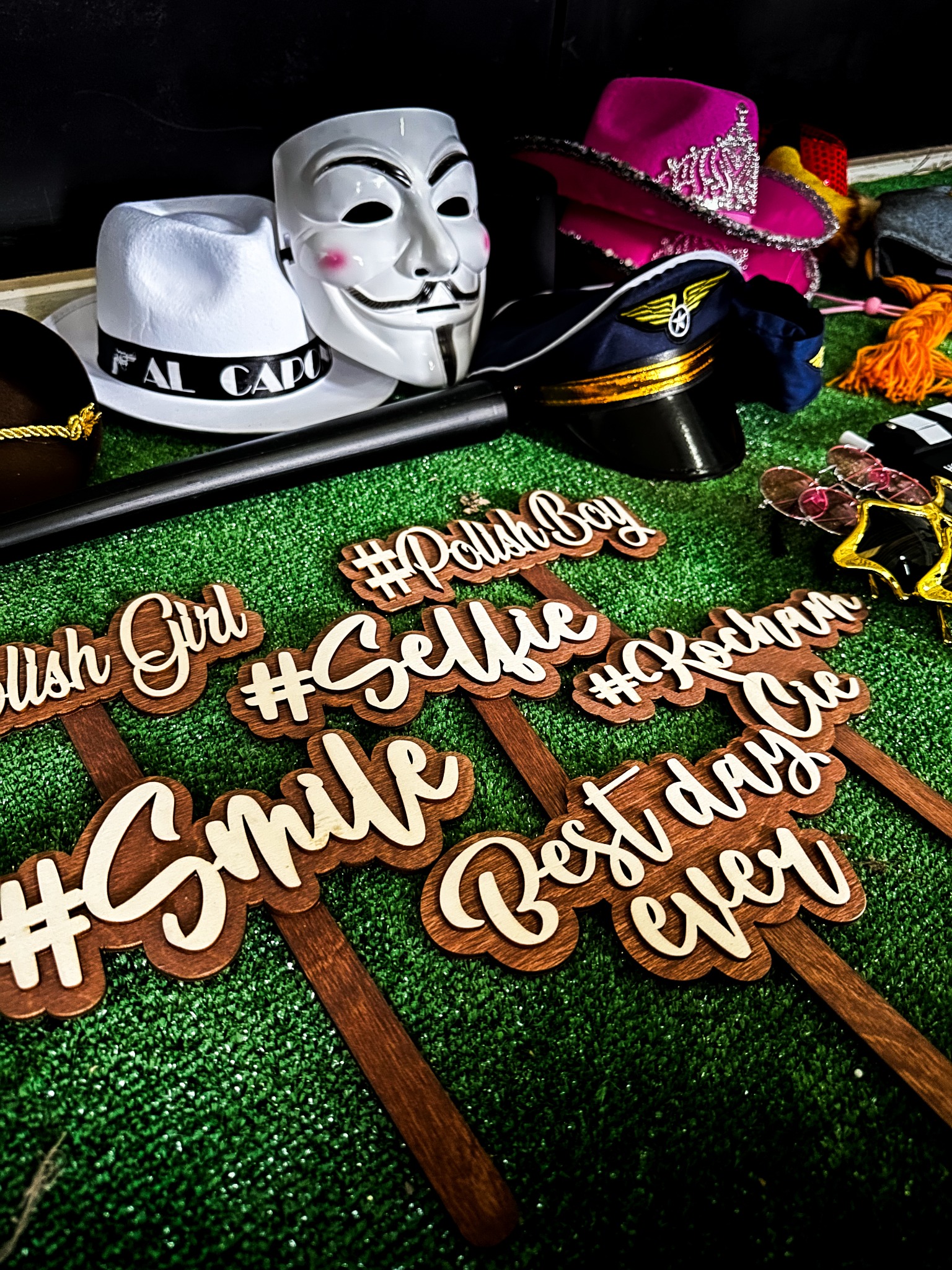 Różnorodne rekwizyty do fotobudki: maska, kapelusze, okulary i drewniane tabliczki z napisami #Selfie, #Smile, Best day ever na zielonym tle.