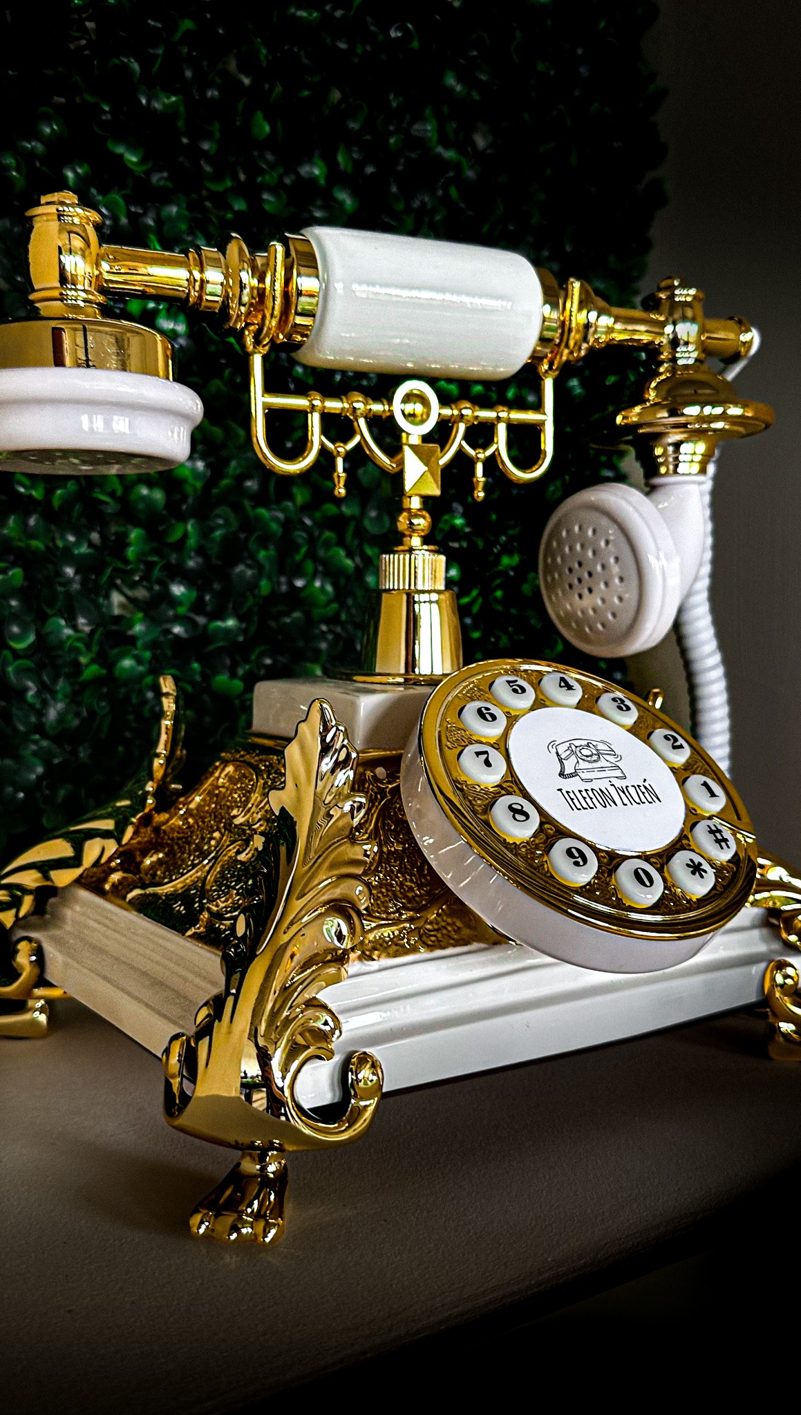 Dekoracyjny, złoto-biały telefon w stylu retro z napisem 'Telefon Życzeń' na tle zielonej ściany roślinnej, element dekoracyjny na imprezę.