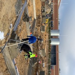 BART-BUD Bartłomiej Kujawa - Dwóch murarzy w trakcie wznoszenia fundamentów z bloczków betonowych. Widoczne narzędzia, wykop i częściowo ukończona konstrukcja. W tle budynek i sprzęt budowlany.