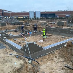 BART-BUD Bartłomiej Kujawa - Wykop pod fundamenty z częściowo wymurowanymi ścianami z szarych bloczków. W tle pracownicy budowlani i budynek handlowy w pochmurny dzień.