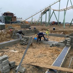 BART-BUD Bartłomiej Kujawa - Budowa fundamentów z bloczków betonowych na placu budowy z atrakcjami parku rozrywki w tle. Dwóch pracowników przy pracy, widoczne narzędzia i materiały budowlane.