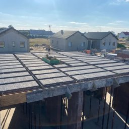 BART-BUD Bartłomiej Kujawa - Wysoki kąt na budowę domu jednorodzinnego z szarym stropem Teriva i zbrojeniem. W tle dwa ukończone domy w stanie surowym zamkniętym i błękitne niebo.