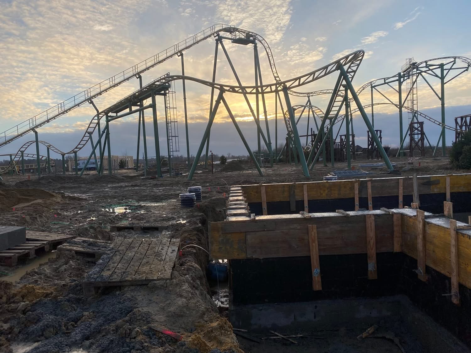 Fundamenty pod konstrukcję kolejki górskiej w parku rozrywki, widoczne drewniane szalunki i zbrojenie, w tle stalowa konstrukcja kolejki i zachmurzone niebo.